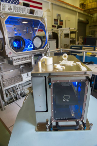 Foreground: Our first 3D printer, contracted by NASA, which launched to the International Space Station on September 21st, 2014. The ivory objects on top are duplicates of what will soon be some of the first objects ever printed off-Earth.  Background: The Microgravity Science Glovebox that will contain the printer during the 3D Printing in Zero-Gravity Experiment.  Image Credit: NASA/Emmett Given